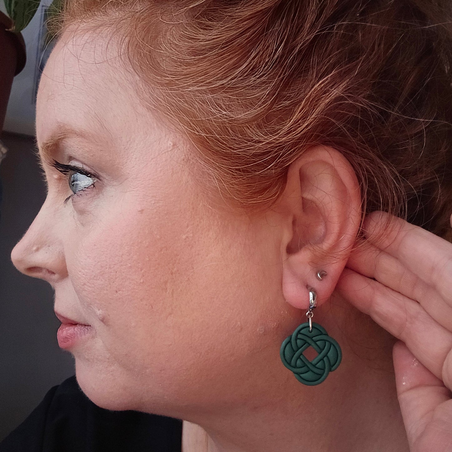 Celtic Knot Dangles by wixies featuring dark green woven knot design on silver hoops worn on model’s ear close-up